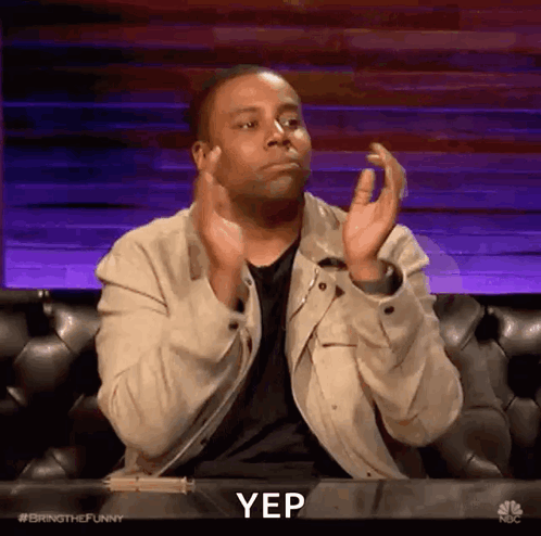 A man seated on a talk-show set claps with a neutral reaction.