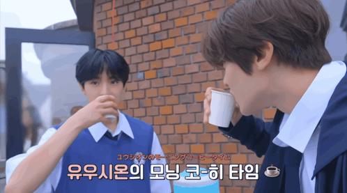 two young men drinking from cups in front of a brick wall with foreign writing