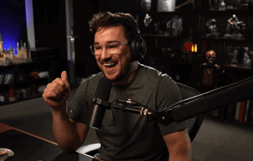 A smiling man at a microphone gives a thumbs up in a dark room.