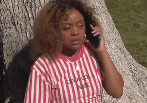 A woman leans against a tree while talking on a phone with a displeased expression.