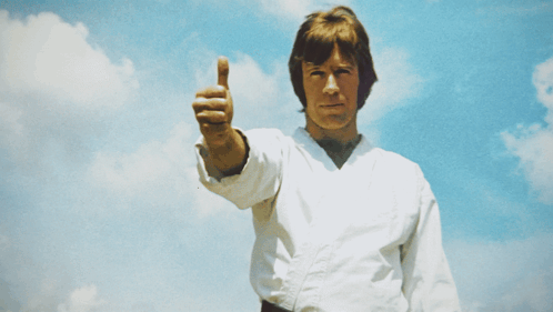 Chuck Norris gives a thumbs up against a bright blue sky.