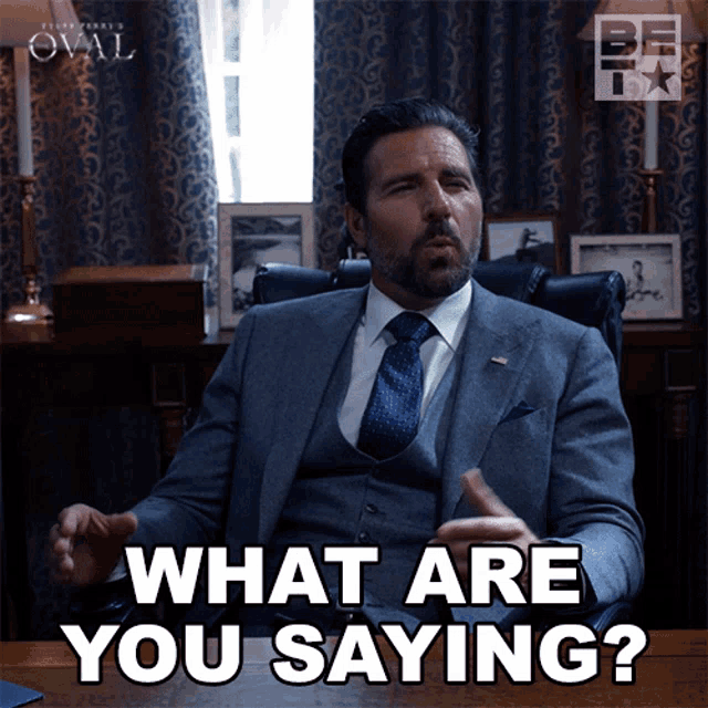 a man in a suit and tie is sitting at a desk and says " what are you saying "