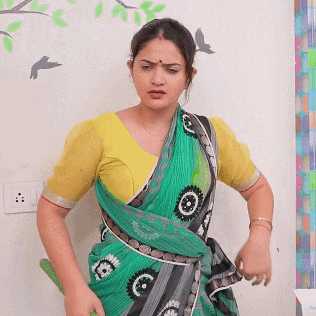 a woman wearing a green saree and a yellow blouse stands in front of a white wall