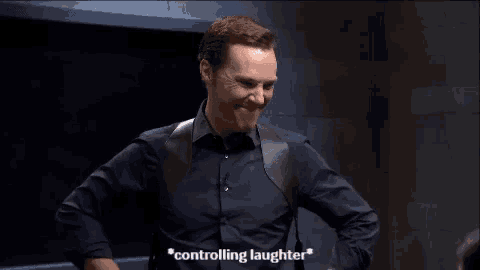 a man in a black shirt is laughing and saying `` controlling laughter '' while standing in a dark room .