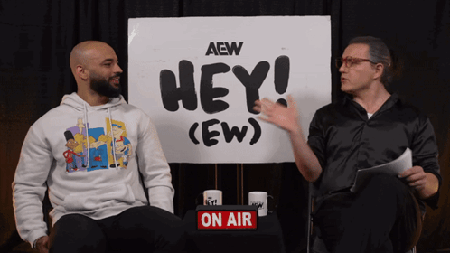 Ricochet and RJ City sit together in front of the “HEY! (EW)” backdrop.