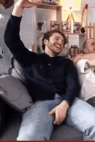 a man is sitting on a couch with his arm in the air and another man is hugging him .