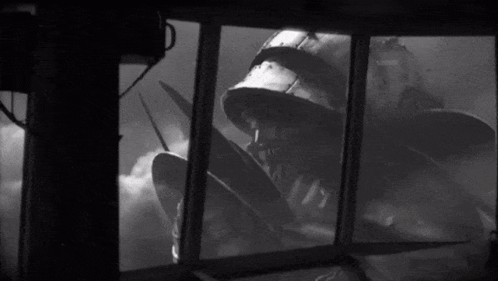 a black and white photo of a man in a helmet looking out a window at a monster .