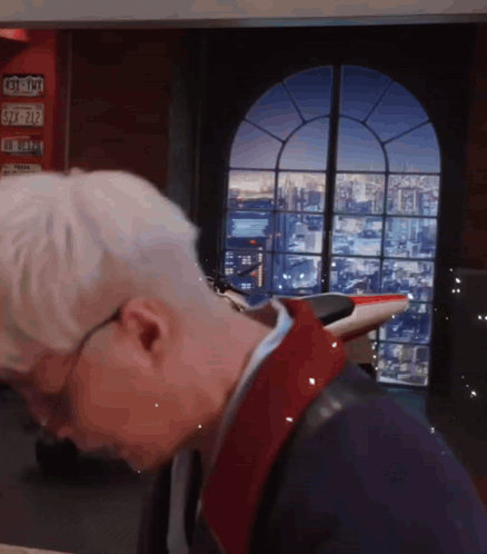 a man with white hair and glasses stands in front of a window with a license plate that says s7 727