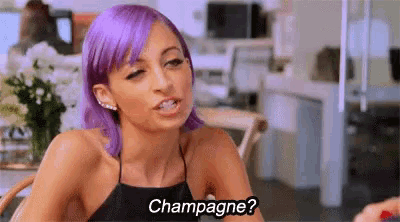 a woman with purple hair is sitting at a table talking to another woman and says `` champagne ? ''