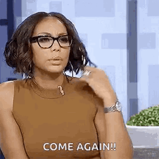 a woman wearing glasses and a brown tank top is sitting in front of a wall and says `` come again '' .