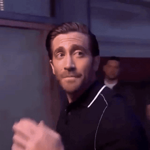 A man in a dark shirt gives a restrained wave-like goodbye gesture.