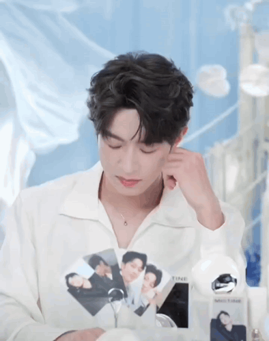 a young man in a white shirt is sitting at a table with his eyes closed and holding a picture of himself .